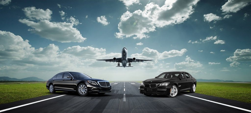 Two black luxury sedans on a runway as a passenger jet takes off overhead